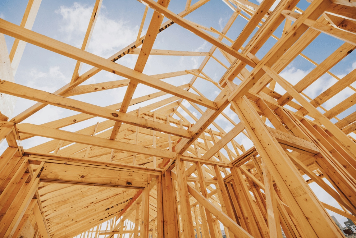 Wooden framework of a house under construction against a blue sky with clouds, showcasing beams and trusses.