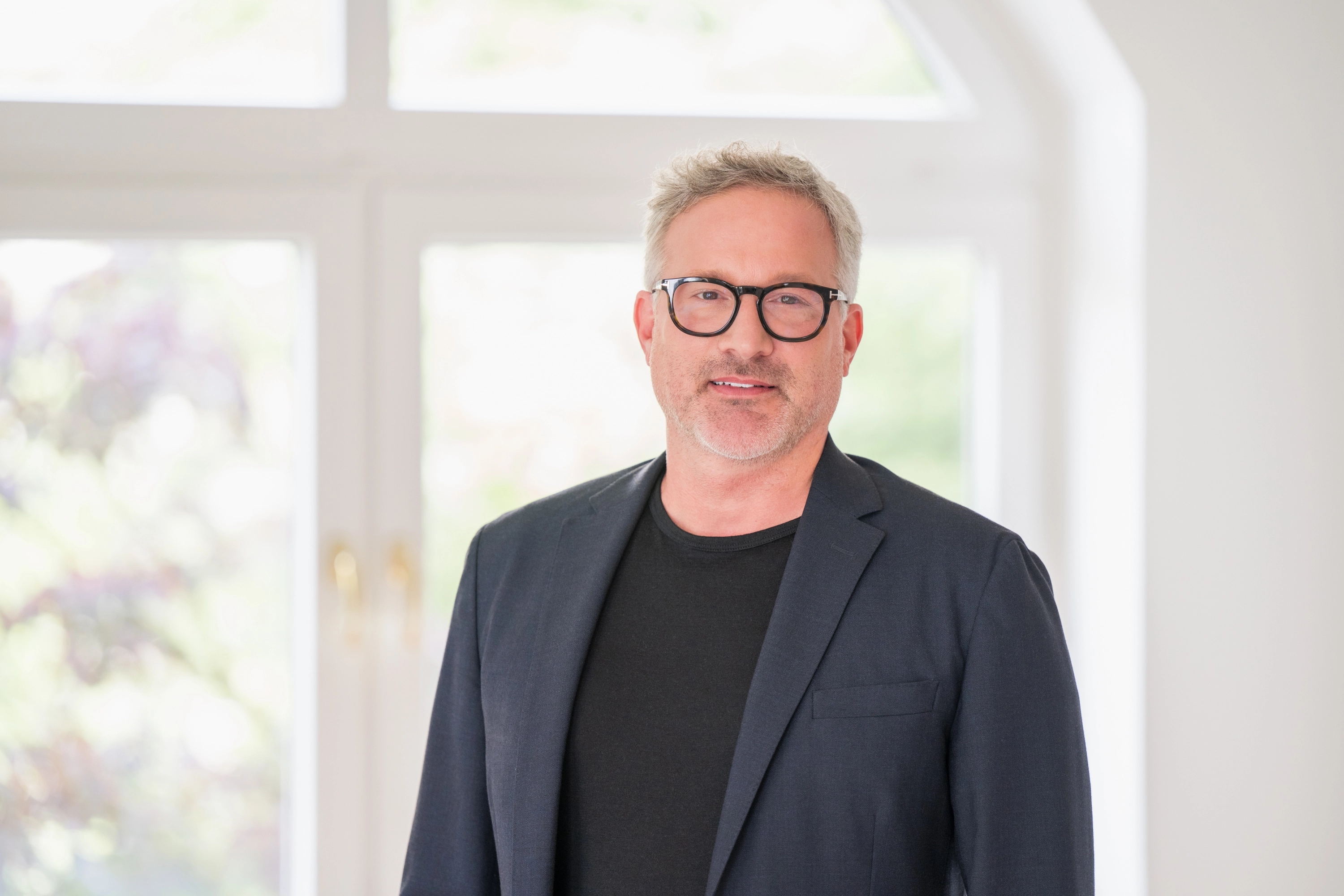 Middle-aged man with gray hair and black glasses in a dark blazer and t-shirt, standing before a bright window.