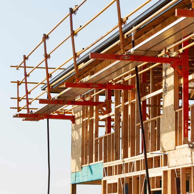 Close-up of a wooden building under construction with red scaffolding and metal beams extending from the structure against a clear sky.