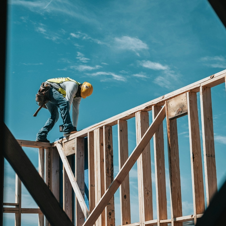 Construction worker in a yellow helmet and safety gear balancing on a wooden frame under a clear blue sky.